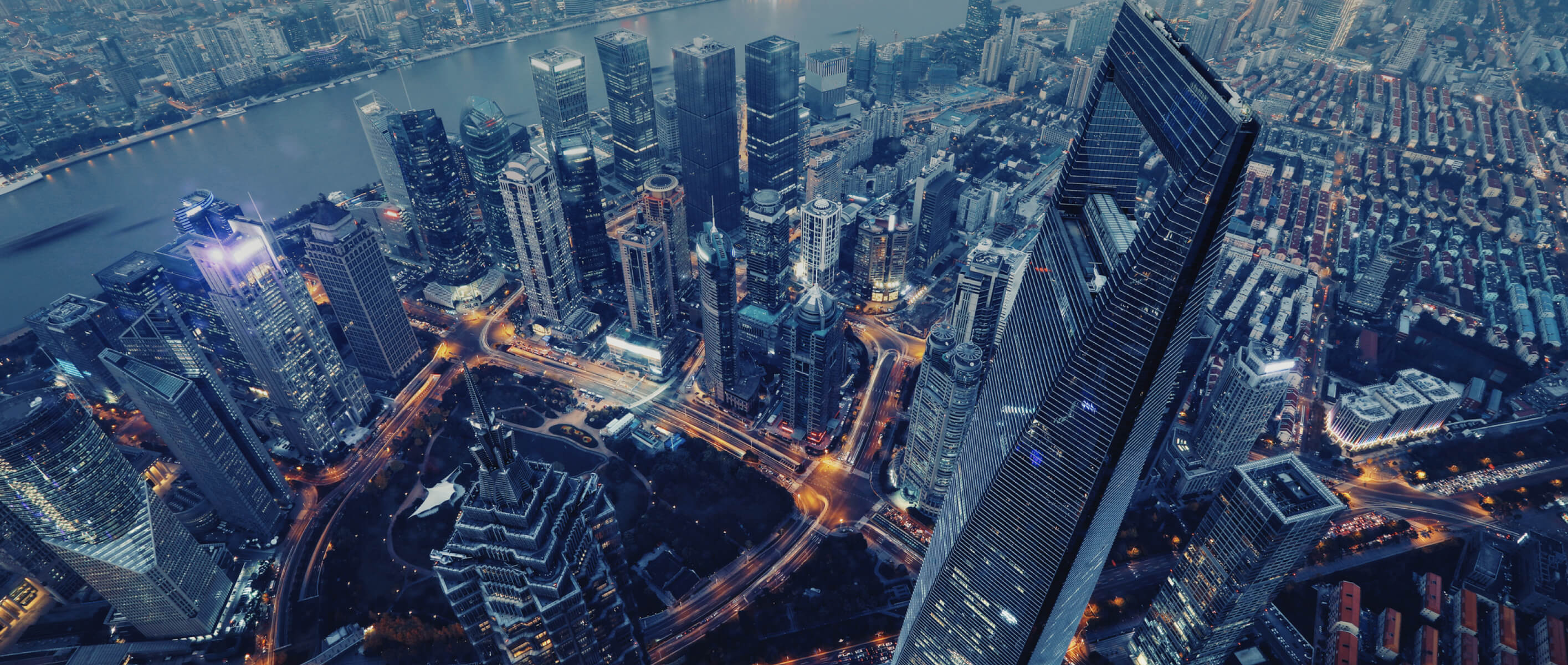 A high-angle aerial view of a Shanghai city skyline at dusk, featuring illuminated skyscrapers, busy roads with moving traffic, and a river in the background.